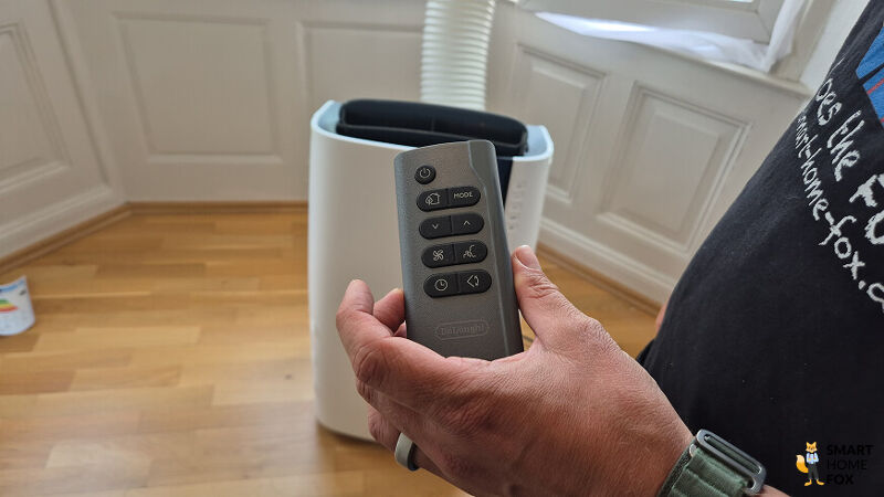 Hand hält Fernbedienung einer mobilen Klimaanlage vor Gerät mit Abluftschlauch im Schlafzimmer